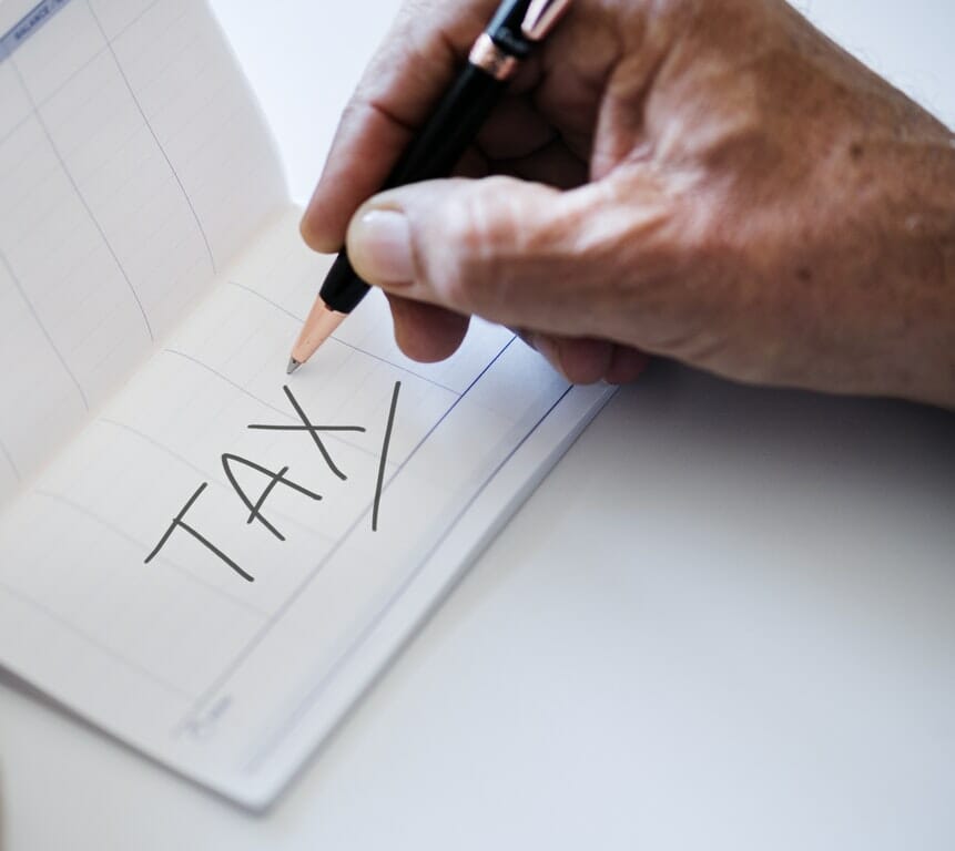 A hand writing the keyword "tax" on a piece of paper.