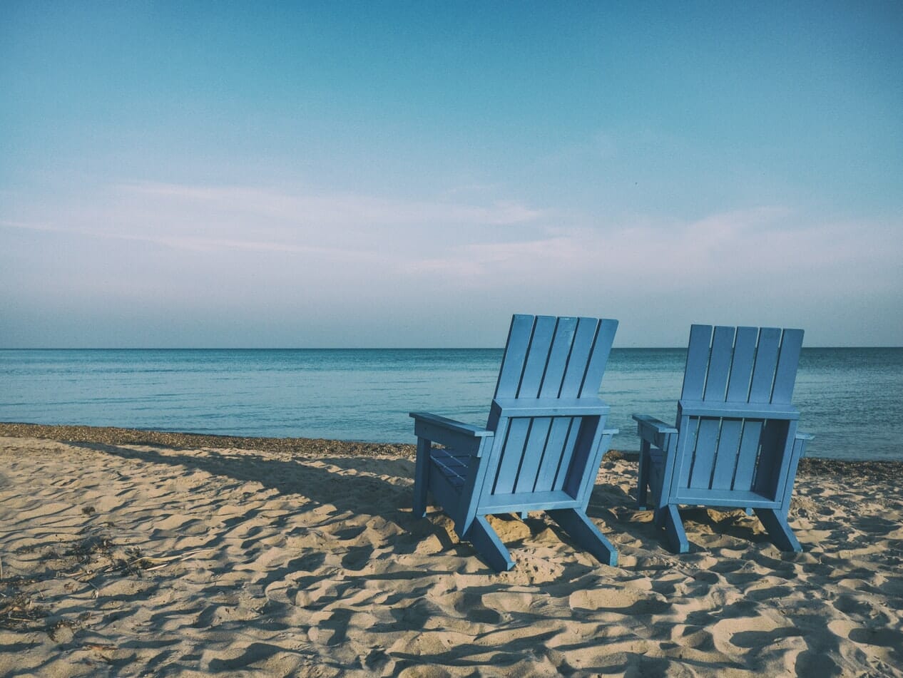 Two blue adirondack chairs on a sandy beach.