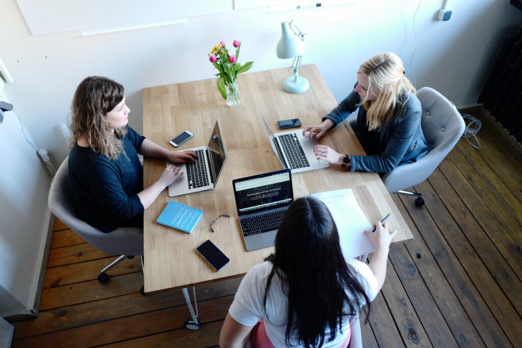 In the accompanying image, three professional businesswomen are depicted engaging in an intense discussion around a table. Each is dressed in smart attire and has a laptop open before them, signifying the importance of technology in modern business. Their focused expressions and collaborative posture convey a strong sense of teamwork and professionalism.