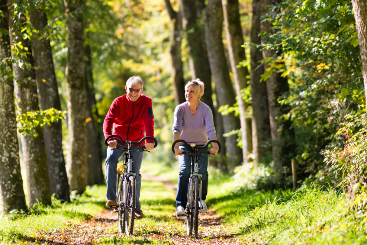 Two older people riding bicycles through a wooded area.