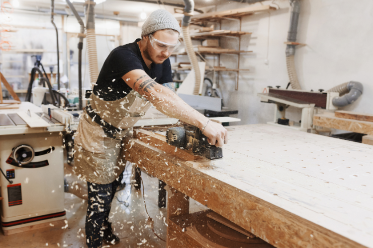 A craftsman in a workshop using a handheld power tool on a large wooden board, with wood shavings flying around.